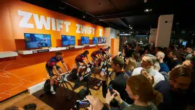 Zwiftのバーチャルサイクリングイベントで、選手たちが固定された自転車で仮想コースを走り、競い合っています。大勢の観客が熱心に見守り、スマートフォンのカメラを向ける姿も見られます。会場は活気に満ちています。