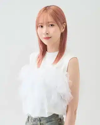 ピンク色の髪の若い女性が白いフリル付きのトップスを着用し、カメラに向かって優しく微笑んでいるポートレート写真