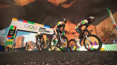 オリンピックバーチャルシリーズの一環として開催されている、砂漠のような仮想空間でのサイクリングレースの様子です。複数のサイクリストがカラフルなユニフォームを着用し、オリンピックのロゴが描かれたアーチを背景に激しく競い合っています。