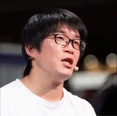 白いTシャツを着て眼鏡をかけたアジア人男性が、ラペルマイクを付けて真剣な表情で話している様子を捉えた画像です。