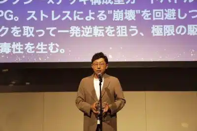 眼鏡をかけた男性がステージ上でマイクを持ち、トロフィーのようなものを手にスピーチをしている