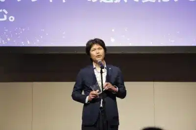 スーツを着た男性がマイクの前でスピーチをしており、手に透明なトロフィーのようなものを持っている