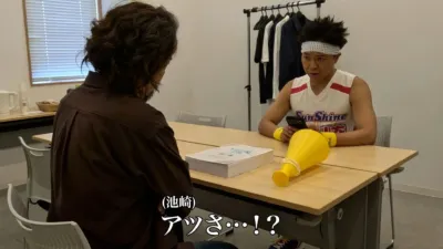 会議室で二人の男性が向かい合って座っています。一人はスポーツウェア姿で驚いた表情をしており、テーブルにはメガホンとスマートフォンが置かれています。