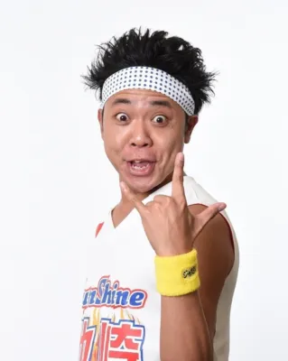 白いヘッドバンドと黄色いリストバンドを身につけた男性が、驚いたような表情でカメラを見つめ、片手でロックサインのポーズをとっています。シャツには「Sunshine」の文字が見えます。