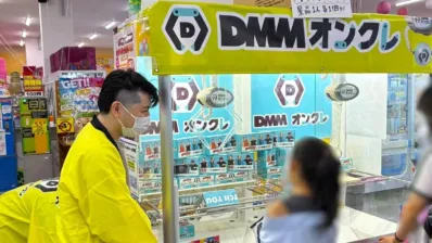 マスクを着用した男性が黄色い法被を着て、DMMオンクレと書かれたクレーンゲームをプレイしている様子