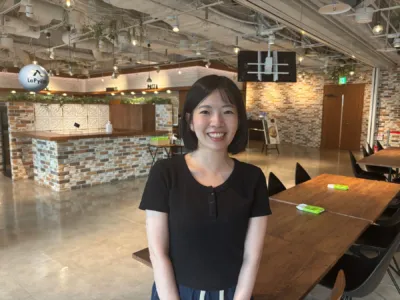 明るい笑顔の若い女性が、モダンな内装のレストランまたはカフェで写っています。店内にはレンガの壁や木製のテーブル、そして「LaPyūta」「NOODLES」「PASTA」の看板が見えます。